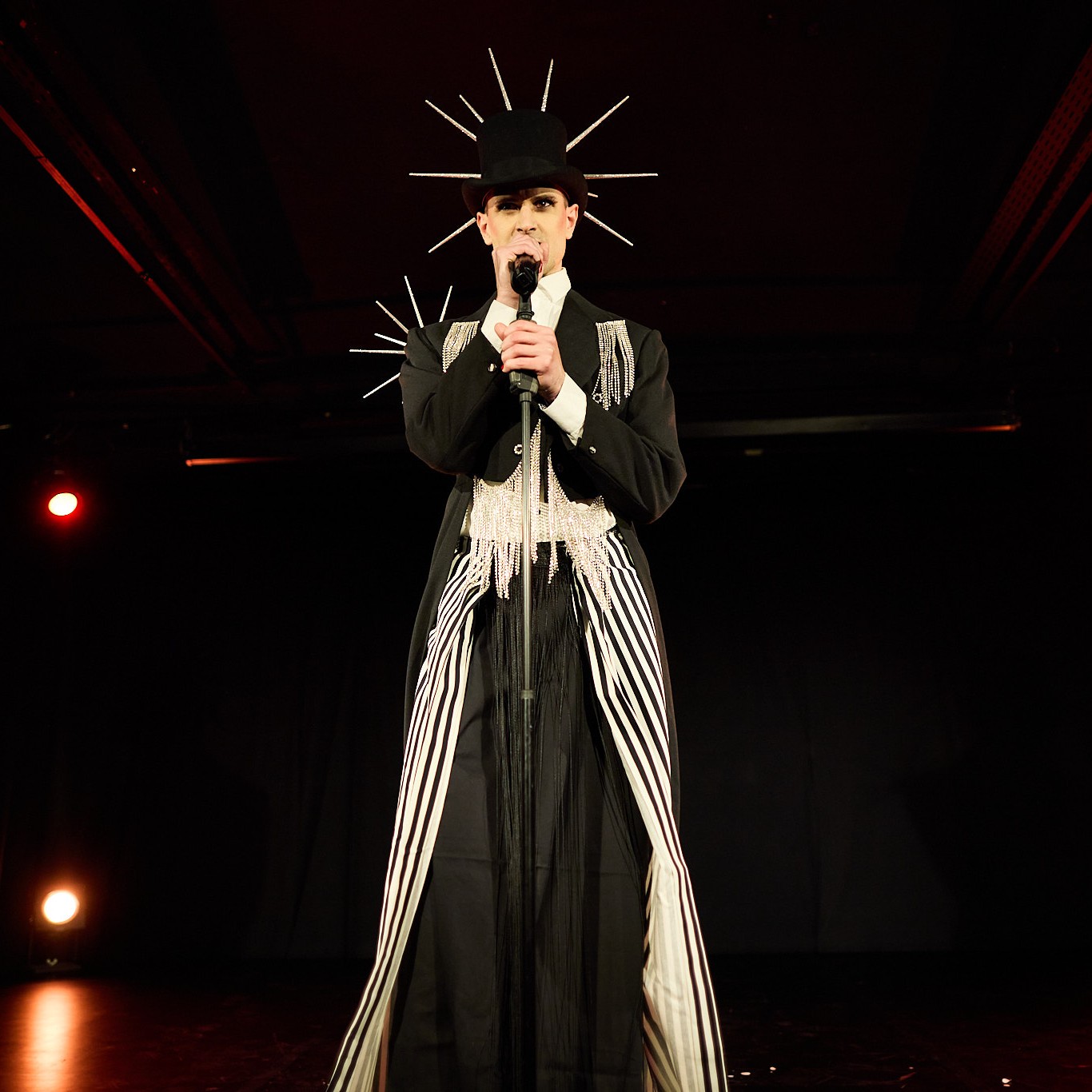 Roylesque is wearing a black top hat with a large silver spiked fascinator, a black tailcoat with sparkly details, a white shirt and a black and white striped skirt.