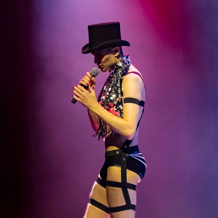 Roylesque is wearing a top hat, a black harness and a sparkly top. They hold a microphone and sing to the audience.
