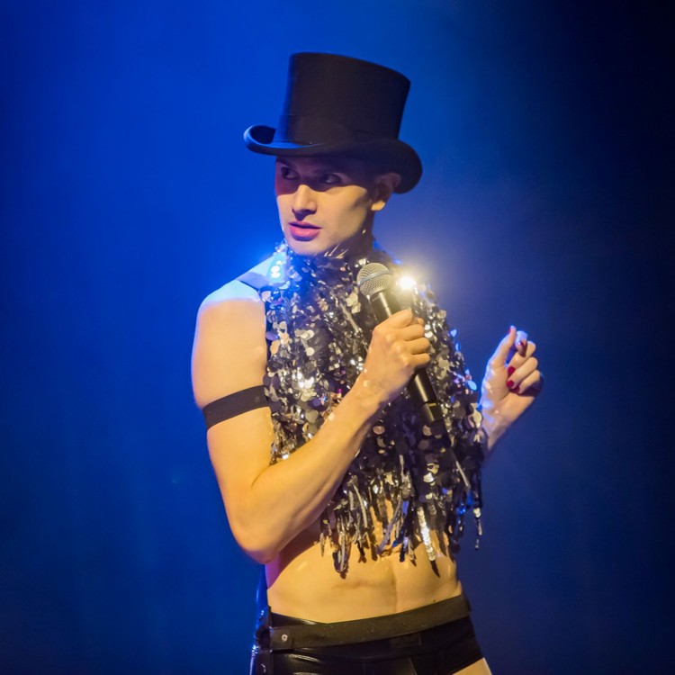 Roylesque is wearing a top hat, a black harness and a sparkly top. There are scars on his ex-posed stomach. He holds a microphone and looks off to the side with an air of nonchalance.