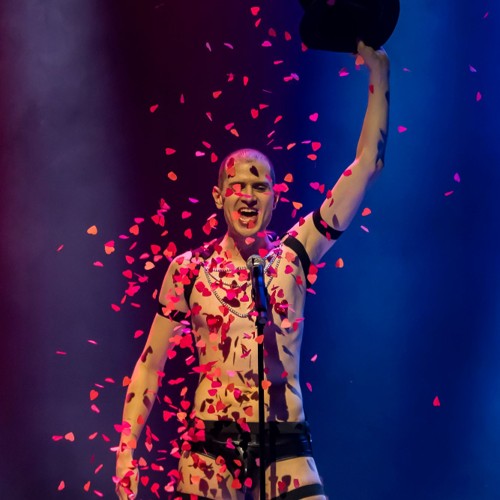 An abundance of red heart-shaped confetti rains down from Roylesque's top hat, which they raise in a powerful pose.
