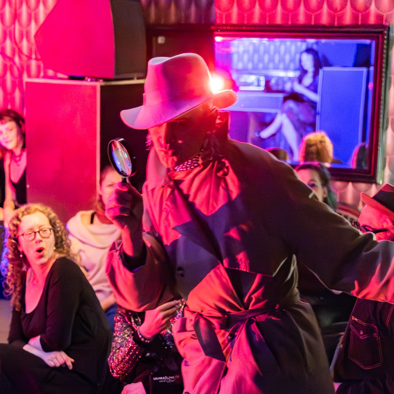 Roylesque in the audience, dressed as a cheeky inspector wearing a hat and a large trench coat, holding a magnifying glass close to his eye.