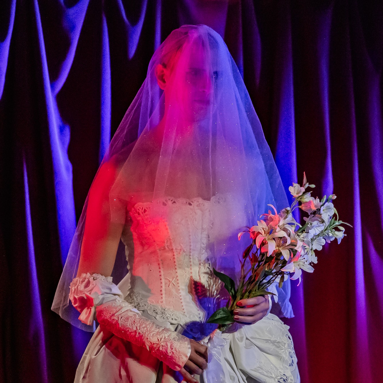 Roylesque wearing an 80s-style wedding dress, with a white glittery veil, an embroidered corset, embroidered gloves with bows, holding a large bouquet of white flowers.