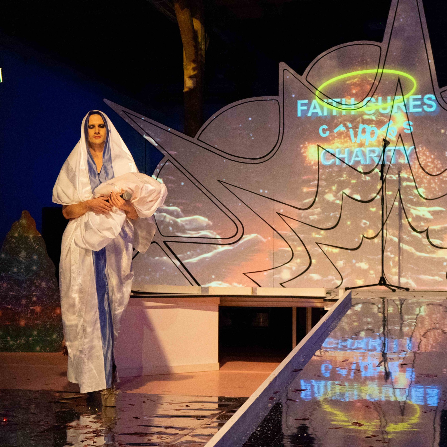 Roylesque is walking onto stage, wearing a white dress with blue detailing and carrying what appears to be a baby wrapped in white cloth.