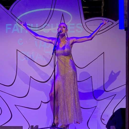 Roylesque is triumphantly standing on stage in a tight golden gown, raising his arms and singing into a microphone.