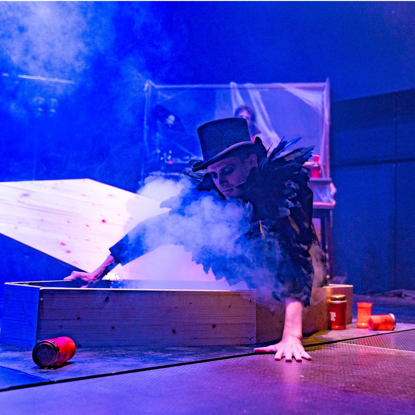 Roylesque is climbing out of a coffin, wearing a black top hat, dark make-up and a large black feathered top.