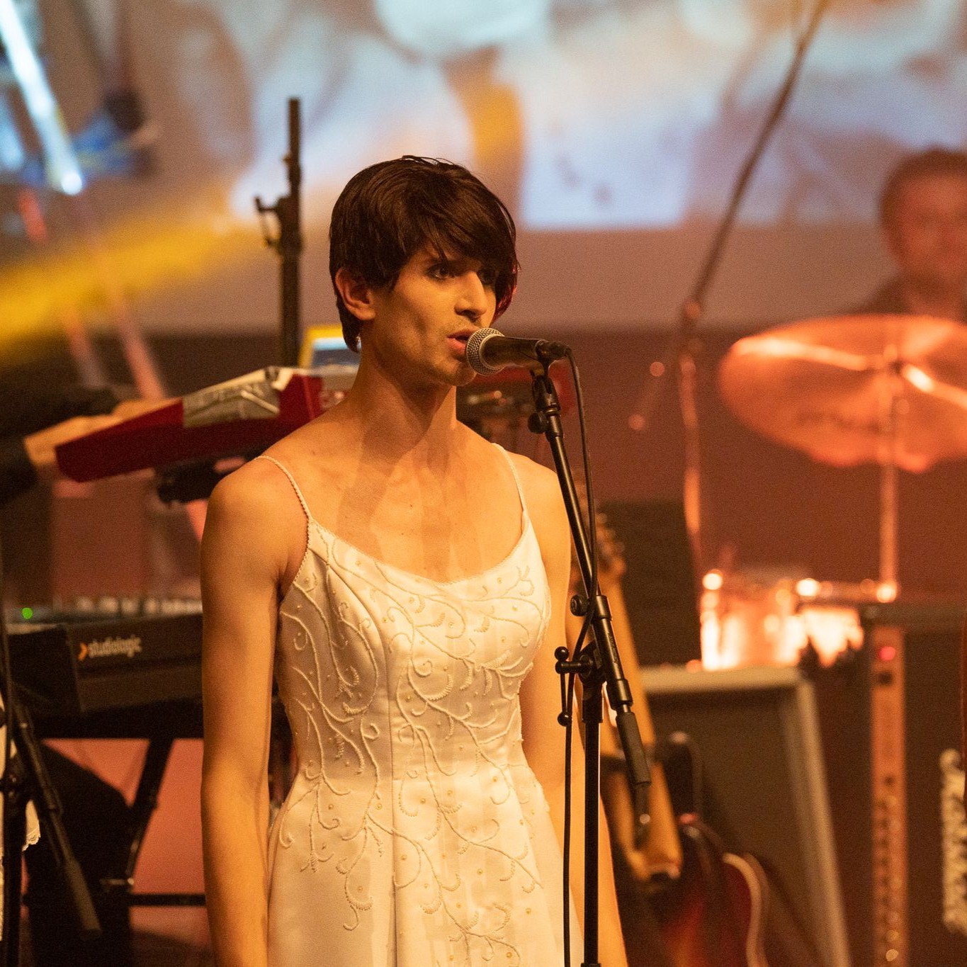 Roylesque is wearing a simple white wedding dress with white embroidery and medium-length brown hair, singing into a microphone.