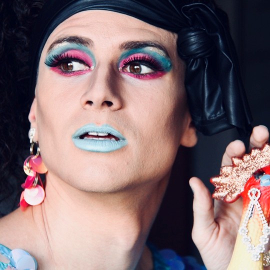 A close-up of Roylesque's face, wearing bright 80's-style makeup in blue and pink, with a black hair band, pink sequined earrings and a blue sequined dress.