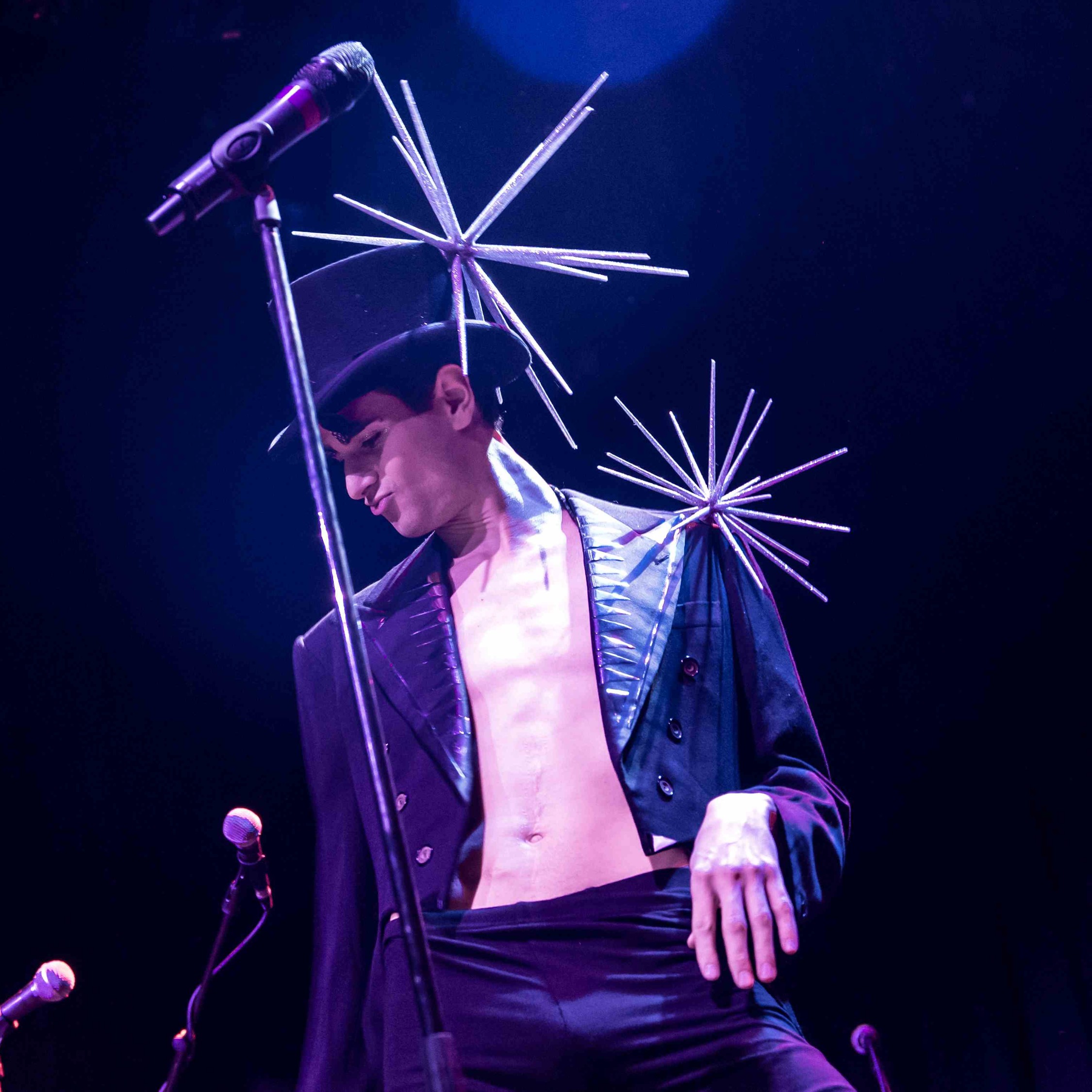 Roylesque is wearing a black top hat with a large silver spiked fascinator, a tailcoat with silver details and black trousers, their stomach is exposed.