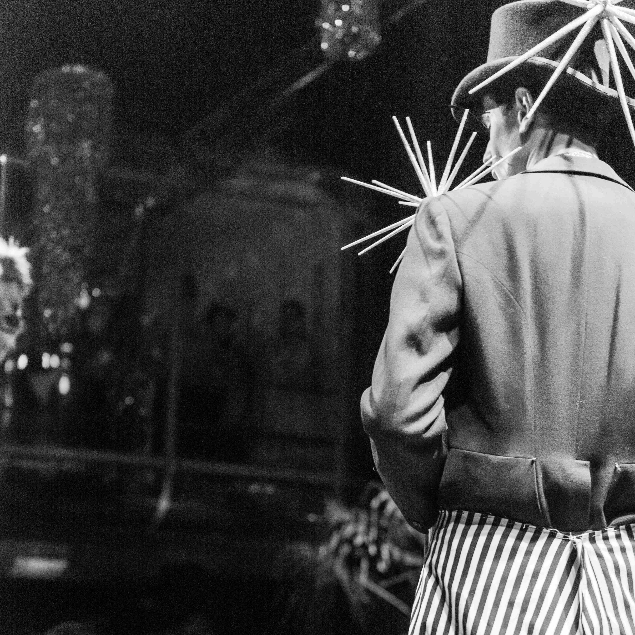 Roylesque is seen from behind as they are looking into the venue, wearing a large silver spiked fascinator on their black top hat, a black top and striped skirt.