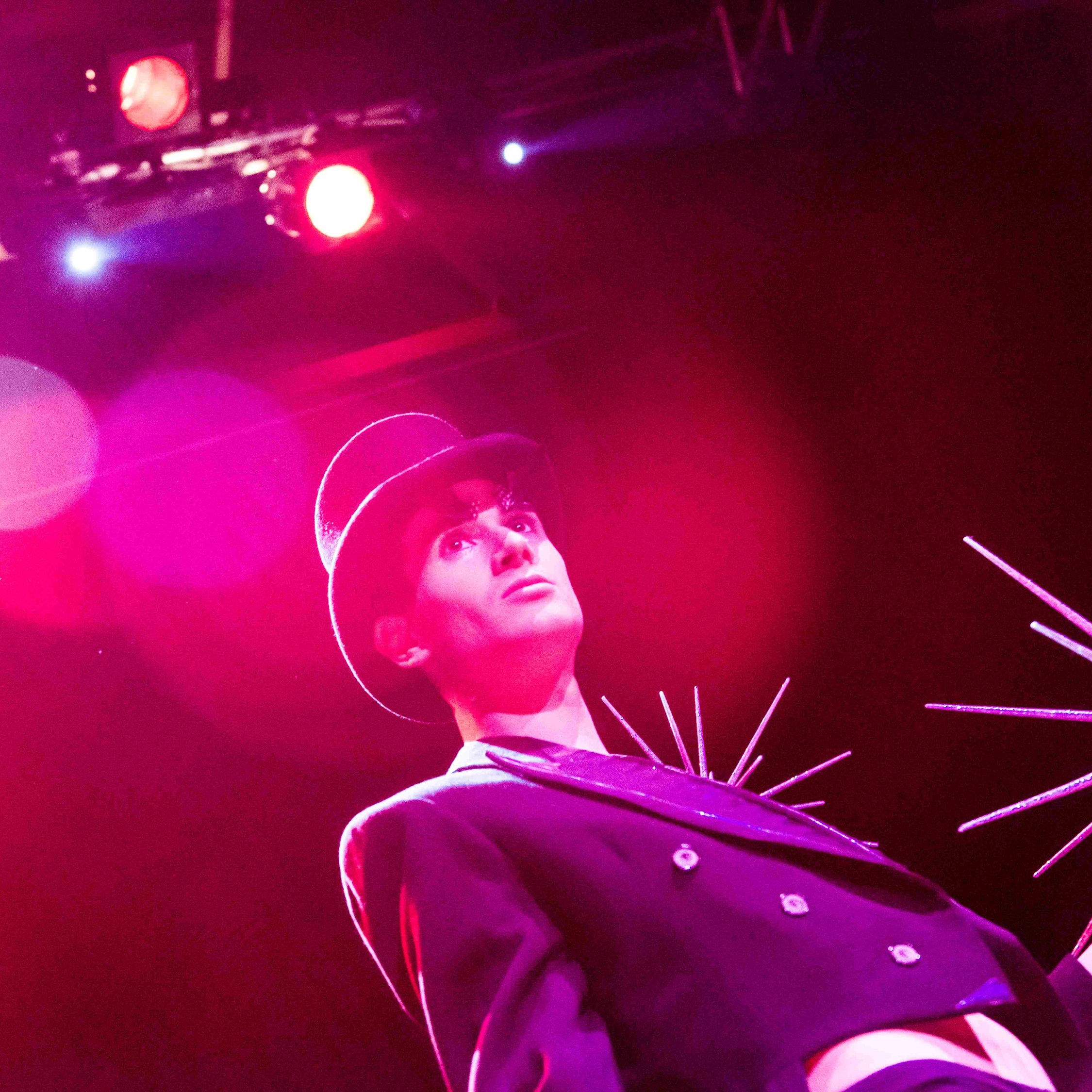 Roylesque has just removed the silver spiked fascinator, which they now hold in their hand, they are wearing a black top hat, black tailcoat with silver detailing, their stomach is exposed. There is a dramatic flash of pink light shining on their face.